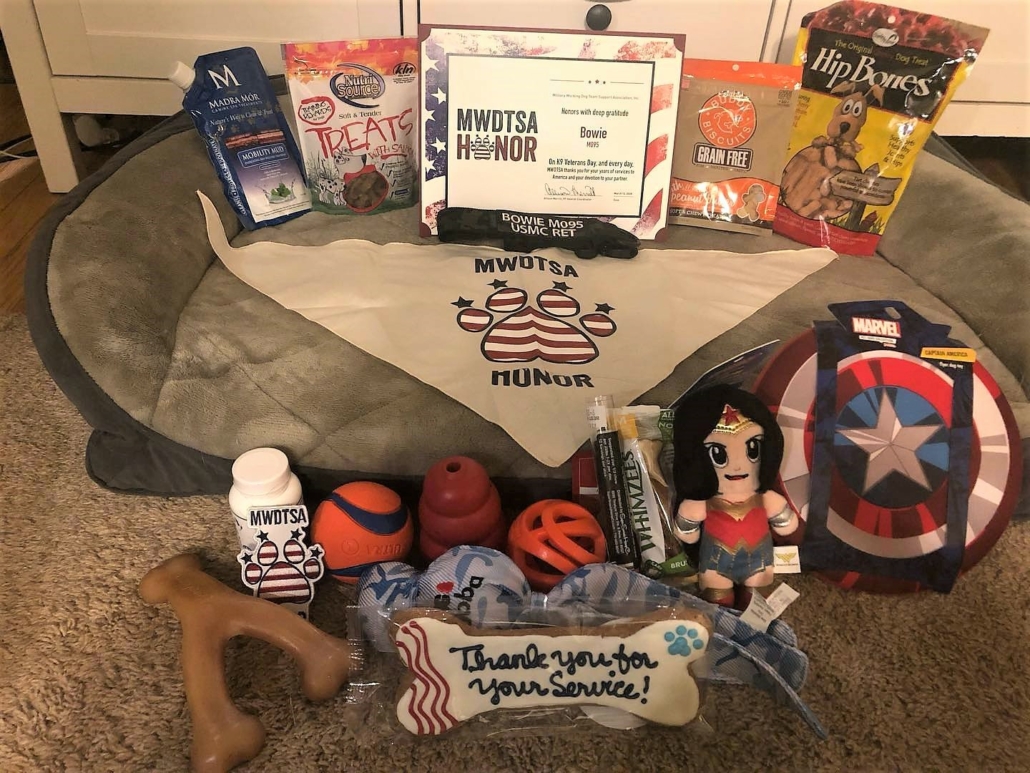 This image shows Honor Box contents from a prior year. In addition to dog treats and toys, RMWDs received a commemorative certificate, dog bandana and dog cookie in the shape of a bone, which says, "Thank you for your service."