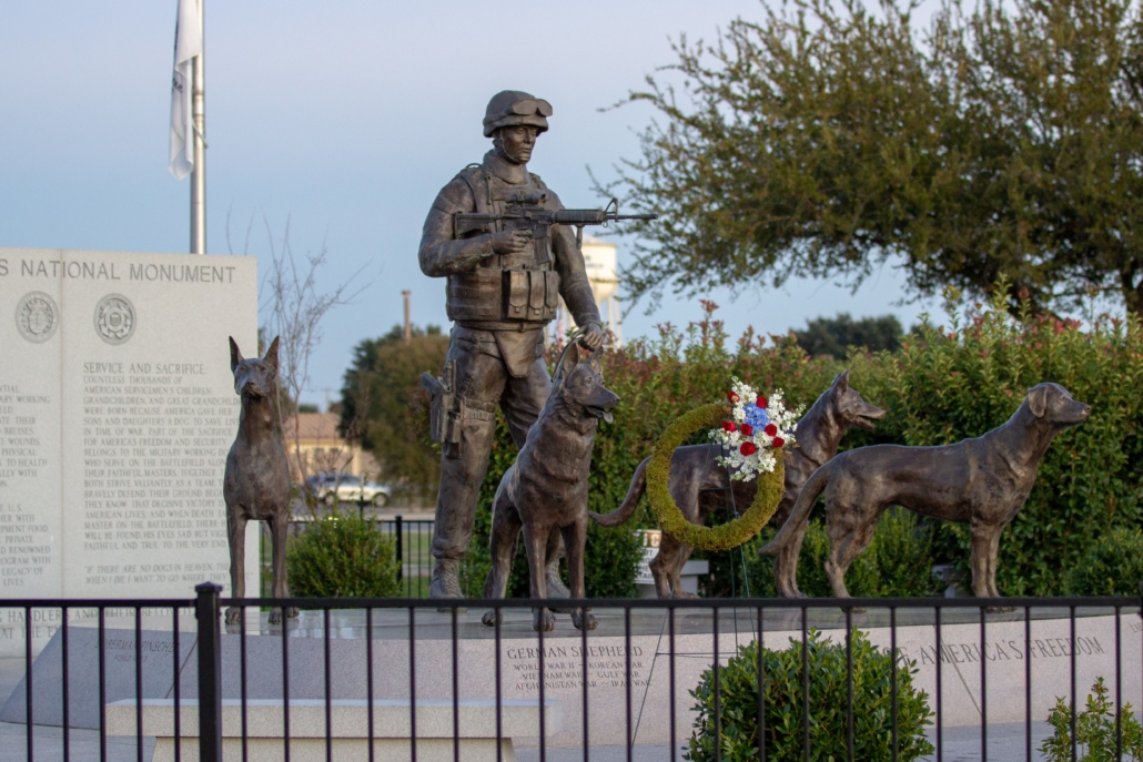 This is a 2019 photo of Military Working Dogs National Monument at Lackland Air Force Base in San Antonio, Texas. A bronze sculpture of a handler stands with his gun ready. Arrayed before him are bronze sculptures of four military working dogs representing the four main breeds used by the military. MWDTSA provided a memorial wreath, displayed on the monument. MWDTSA is providing a wreath for Memorial Day 2020, as well.