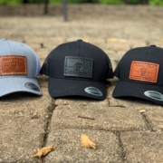This photo shows the MWDTSA FlexFit Snapback hat in three color variations: Charcoal with a brown patch, Black with a brown patch, and Black with a black patch.