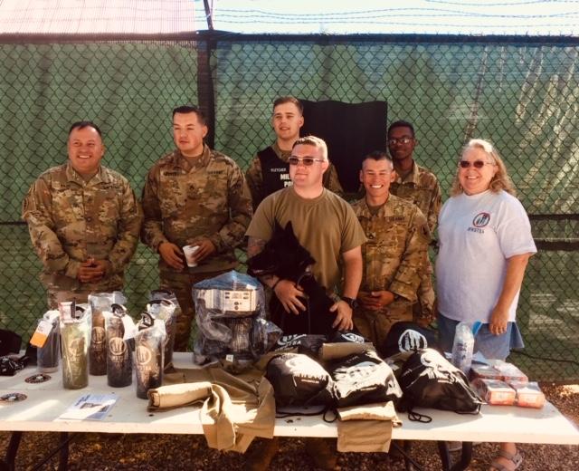 This photo shows the Fort Huachuca handlers and MWDTSA volunteer Linda Costa-Bryan standing behind a picnic table loaded with MWDTSA gifts, including backpacks, FIFTY/FIFTY bottles, t-shirts, and more.