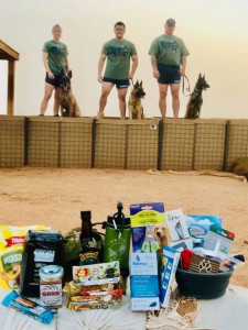 Care package contents sit on a sand bag in the foreground. Three handlers and their MWDs stand in the background modeling the 'Merica-themed MWDTSA t-shirts and MWDTSA athletic shorts that were included in the Q2-2019 care packages.