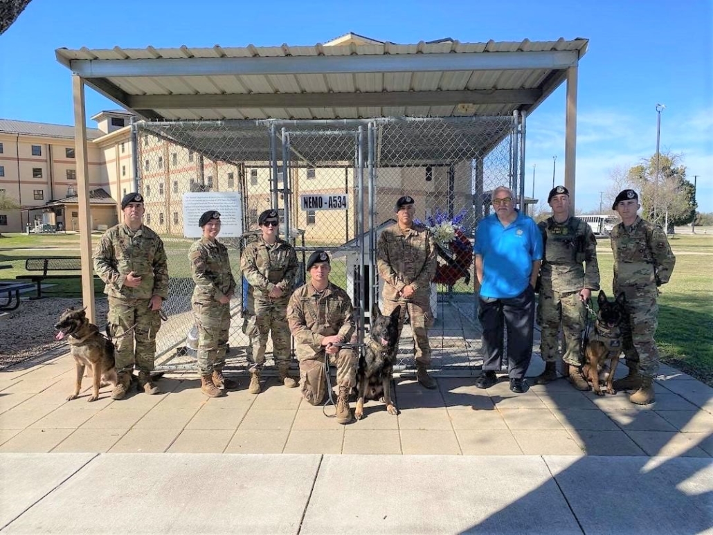 This photo shows handlers and their dogs, along with Security Forces Museum Director Ken Neal, standing in front of Nemo's Memorial at Lackland Air Force Base. In the background is a wreath donated by MWDTSA in honor of Vietnam veteran dog handler Bob Throneburg.
