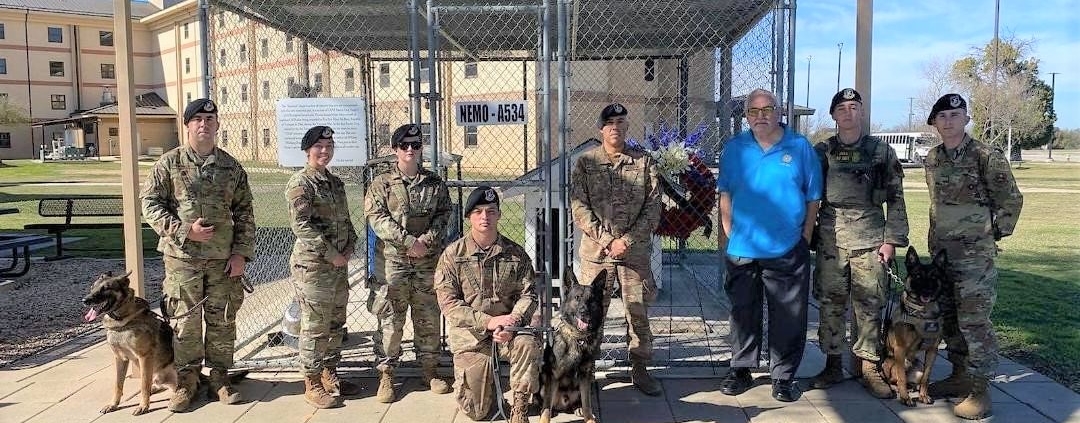This photo shows handlers and their dogs, along with Security Forces Museum Director Ken Neal, standing in front of Nemo's Memorial at Lackland Air Force Base. In the background is a wreath donated by MWDTSA in honor of Vietnam veteran dog handler Bob Throneburg.