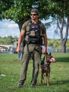 This image shows Justin and Pepper from the TV show SEAL Team. PIctured: Justin is standing beside Pepper, who is wearing a fierce-looking muzzle. Pepper invites you to take part in Pepper's Donation Drop, a holiday toy drive in collaboration with MWDTSA to support U.S. military working dogs.